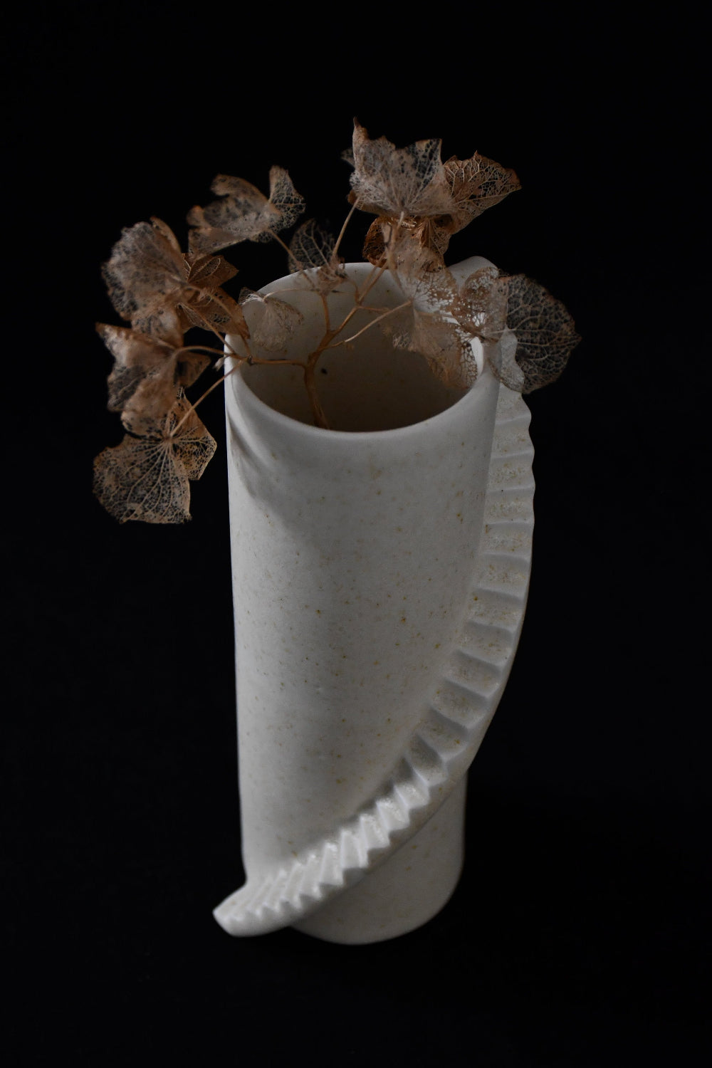 A white porcelain vase with a unique spiral stairs design, featuring dried leaves inside, against a dark background.