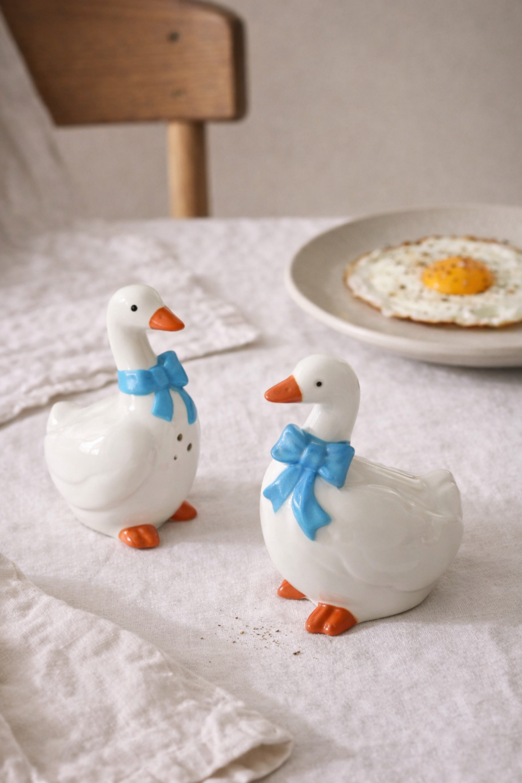 Two ceramic goose figurines with blue bows on a table with a plate of food in the background.