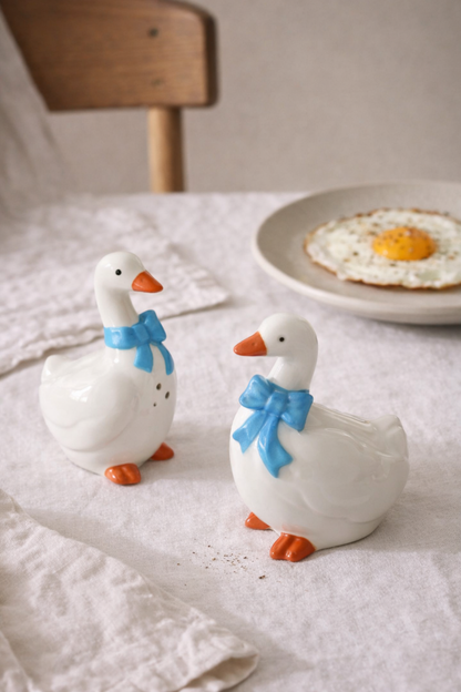 Two ceramic goose figurines with blue bows on a table with a plate of food in the background.