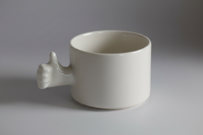 A white porcelain coffee cup with a thumb up gesture as its handle, isolated on a black background.