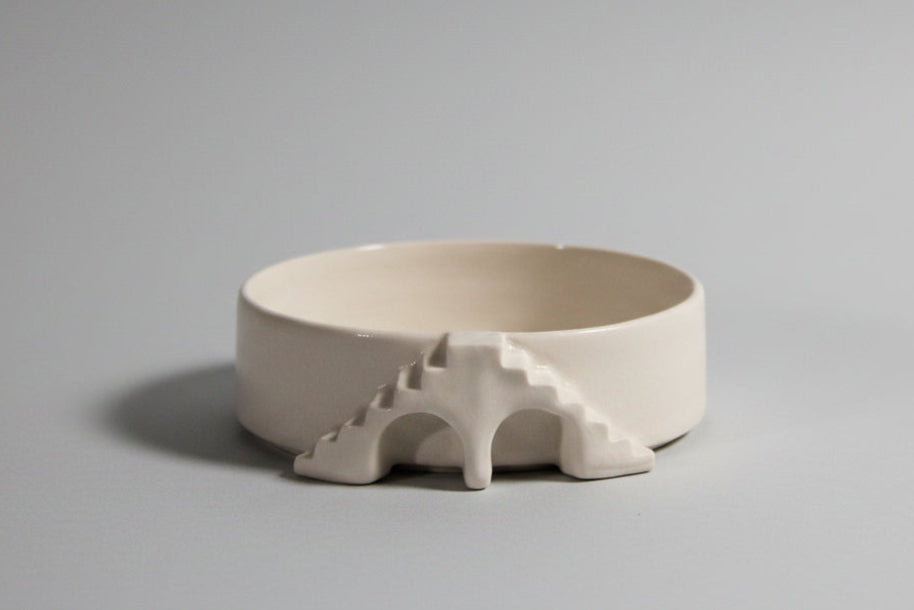 A minimalist off-white ceramic bowl with a sculptural, geometric handle resembling a stylized bridge or arch, set against a solid black background.